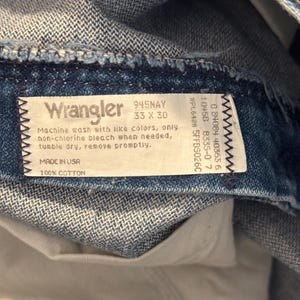 Puede incluir: Una etiqueta de mezclilla azul Wrangler con texto blanco. La etiqueta dice "Wrangler 945NAY 33 x 30" e incluye instrucciones de cuidado. La etiqueta tambi&eacute;n dice "Made in USA 100% Cotton".