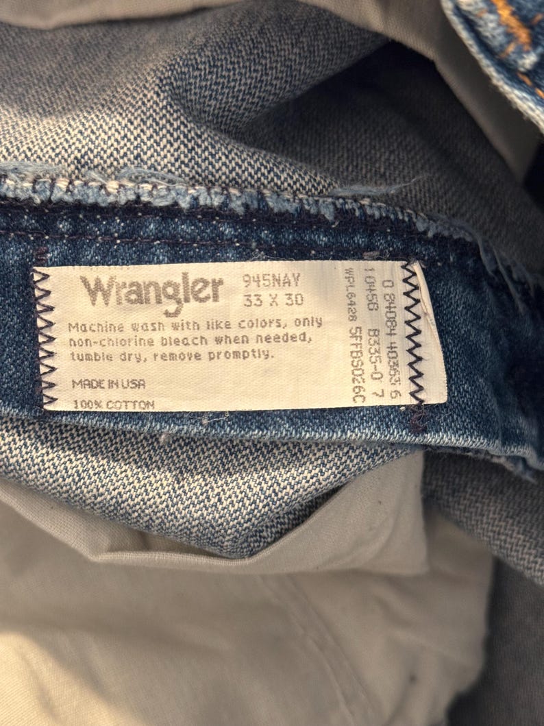Puede incluir: Una etiqueta de mezclilla azul Wrangler con texto blanco. La etiqueta dice "Wrangler 945NAY 33 x 30" e incluye instrucciones de cuidado. La etiqueta tambi&eacute;n dice "Made in USA 100% Cotton".