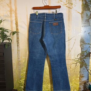 Puede incluir: Un par de pantalones vaqueros azules de pierna acampanada. Los vaqueros tienen un parche de cuero marr&oacute;n en la parte posterior con el logotipo de Wrangler.