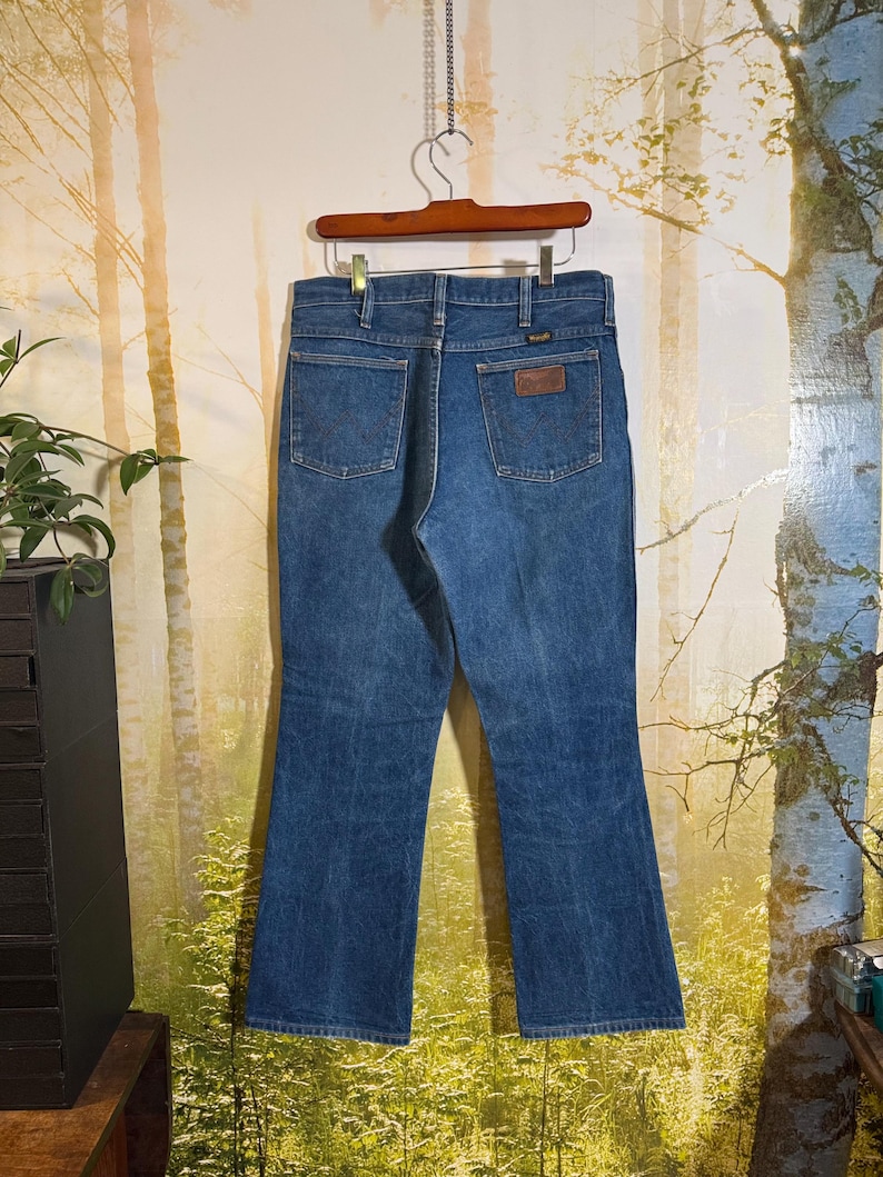 Puede incluir: Un par de pantalones vaqueros azules de pierna acampanada. Los vaqueros tienen un parche de cuero marr&oacute;n en la parte posterior con el logotipo de Wrangler.