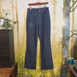 May include: A pair of dark blue denim jeans with a wide leg, hanging on a wooden hanger against a backdrop of a forest scene.