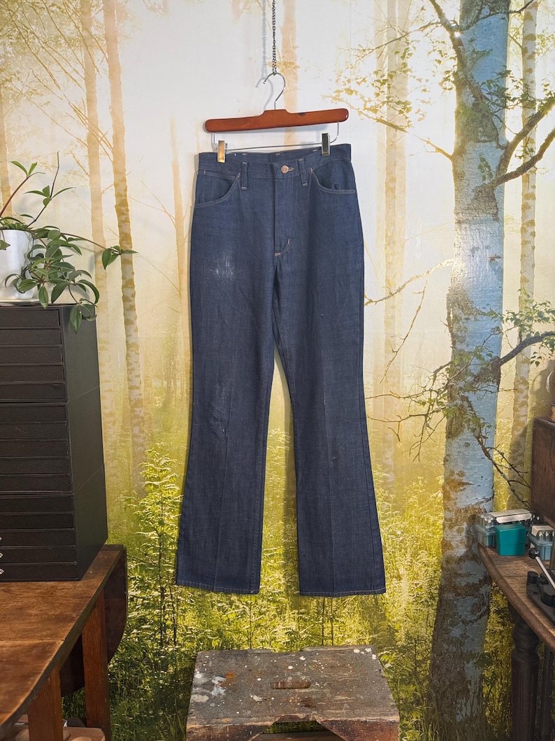 May include: A pair of dark blue denim jeans with a wide leg, hanging on a wooden hanger against a backdrop of a forest scene.