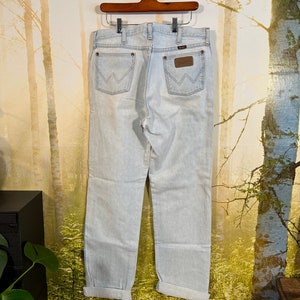 May include: Light blue denim jeans with a cuffed hem. The jeans have a brown leather patch with the Wrangler logo on the back.