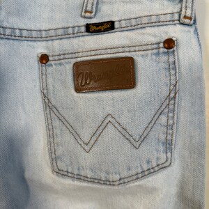 May include: Light blue denim jeans with a brown leather back pocket patch that says "Wrangler" in gold lettering. The jeans have a brown stitching detail around the back pocket.