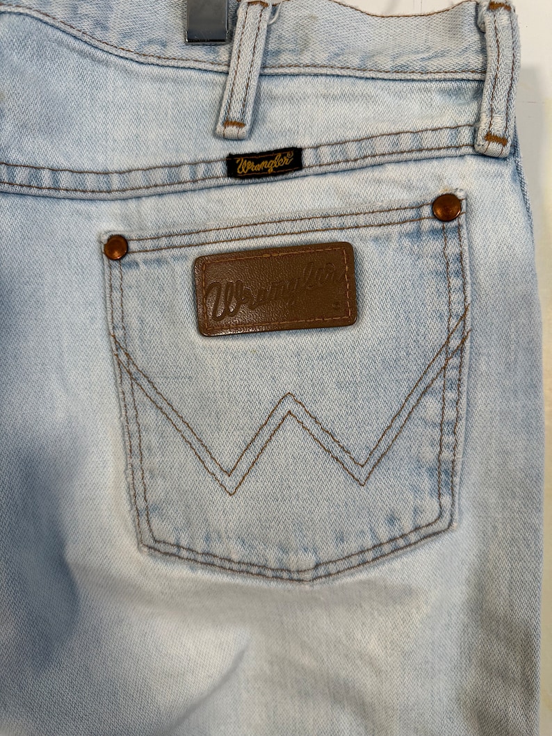 May include: Light blue denim jeans with a brown leather back pocket patch that says "Wrangler" in gold lettering. The jeans have a brown stitching detail around the back pocket.