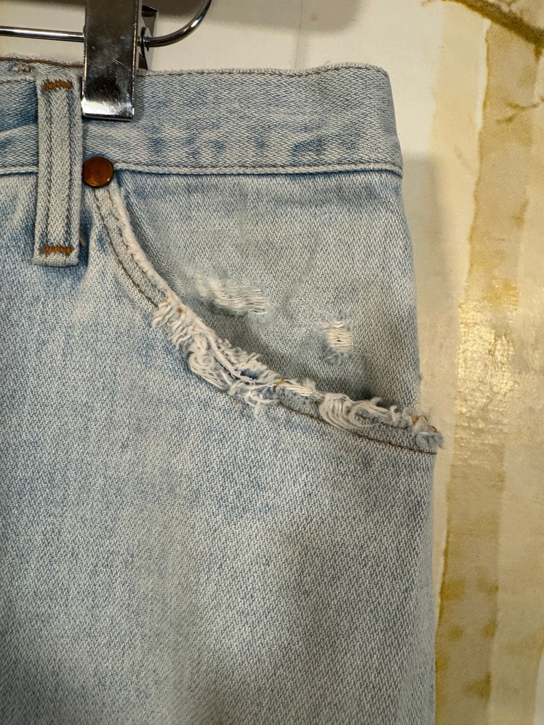 Puede incluir: Vaqueros de mezclilla azul claro con un bolsillo desgastado. El bolsillo tiene un borde deshilachado y un peque&ntilde;o agujero.