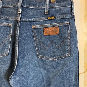 Puede incluir: Un par de pantalones vaqueros azules de mezclilla con un parche de cuero marr&oacute;n en el bolsillo trasero. El parche tiene el logotipo de Wrangler.