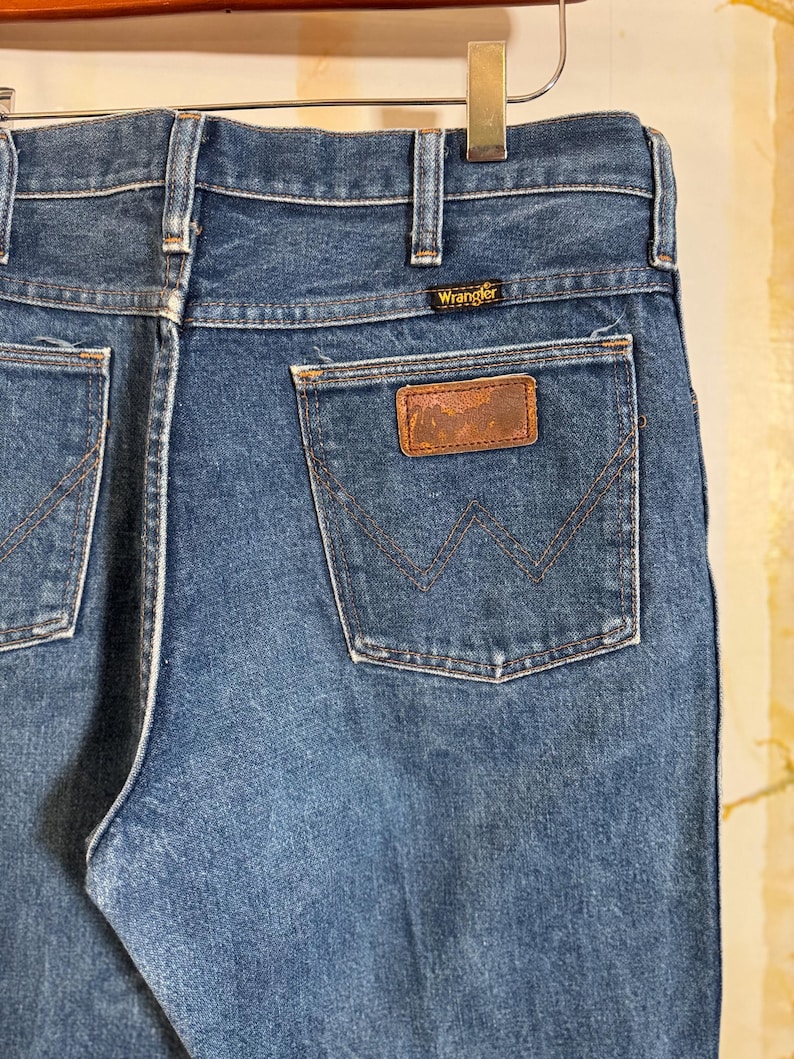 Puede incluir: Un par de pantalones vaqueros azules de mezclilla con un parche de cuero marr&oacute;n en el bolsillo trasero. El parche tiene el logotipo de Wrangler.