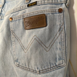 Puede incluir: Vaqueros de mezclilla azul claro con un parche de cuero marr&oacute;n que dice "Wrangler" en el bolsillo trasero.