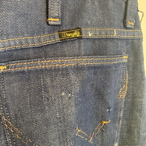 May include: Close-up of the back pocket of a pair of blue denim jeans with a gold Wrangler label. The jeans have a classic style and are made of a durable material.