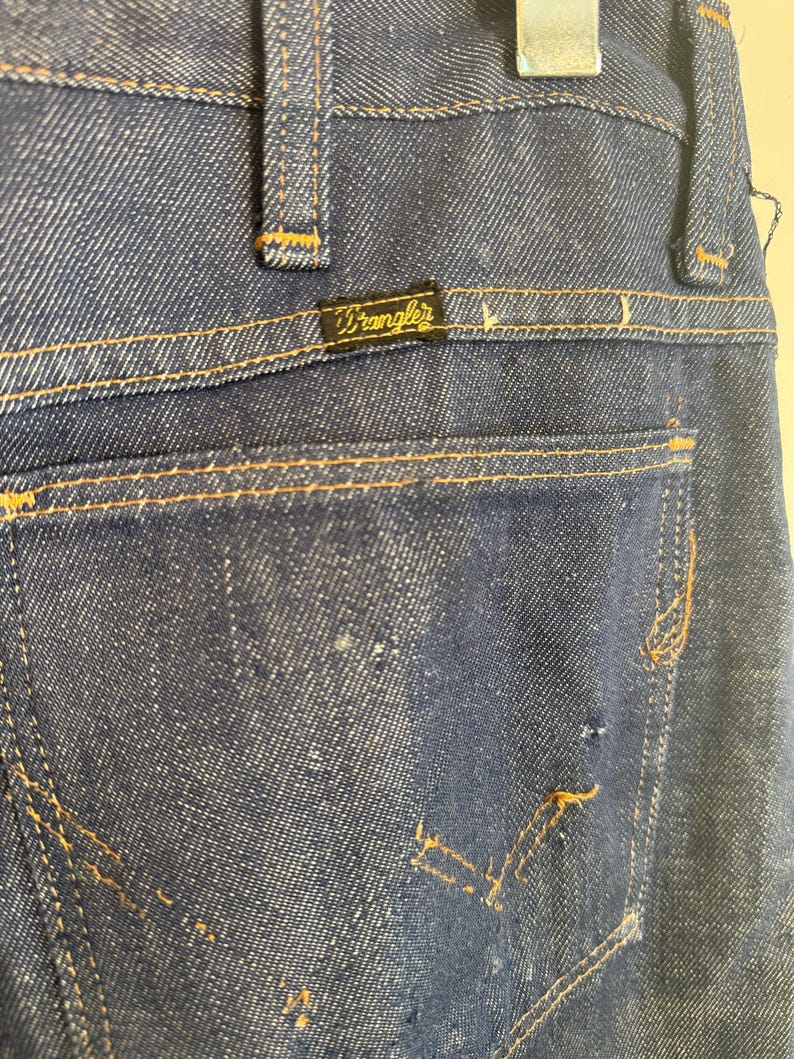 May include: Close-up of the back pocket of a pair of blue denim jeans with a gold Wrangler label. The jeans have a classic style and are made of a durable material.