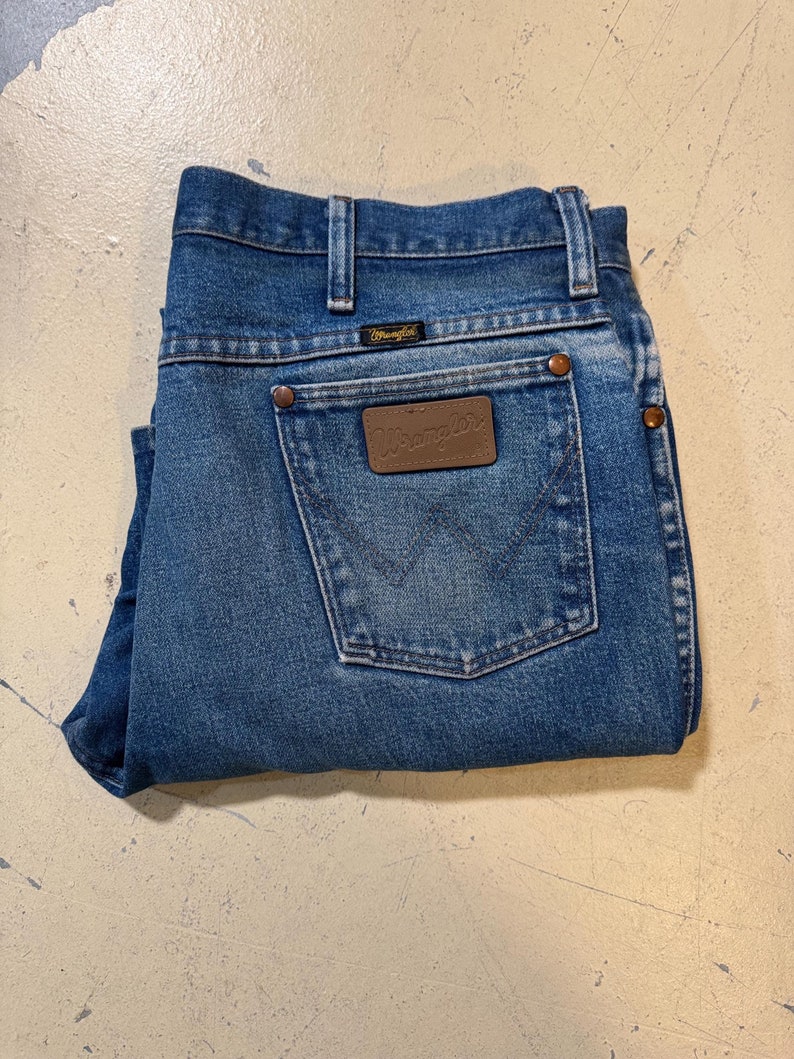 Pu&ograve; includere: Un paio di jeans blu in denim piegati a met&agrave;. I jeans hanno una toppa in pelle marrone sulla tasca posteriore con la scritta "Wrangler" stampata su di essa.