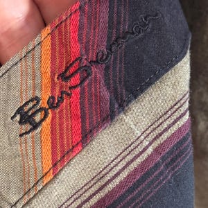 May include: Close-up of a black and brown striped fabric with a red, orange, and black striped label that says "Ben Sherman" in black embroidery.