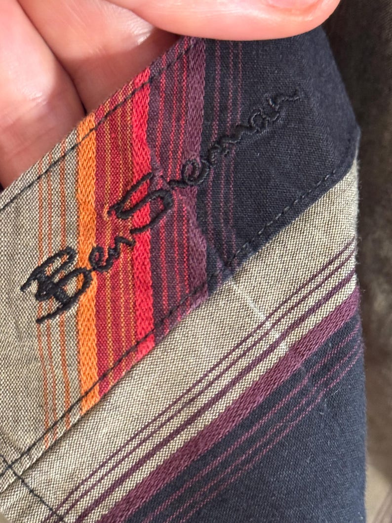 May include: Close-up of a black and brown striped fabric with a red, orange, and black striped label that says "Ben Sherman" in black embroidery.