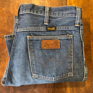 Puede incluir: Un par de pantalones vaqueros azules de mezclilla con un parche de cuero marr&oacute;n que dice "Wrangler" en el bolsillo trasero.