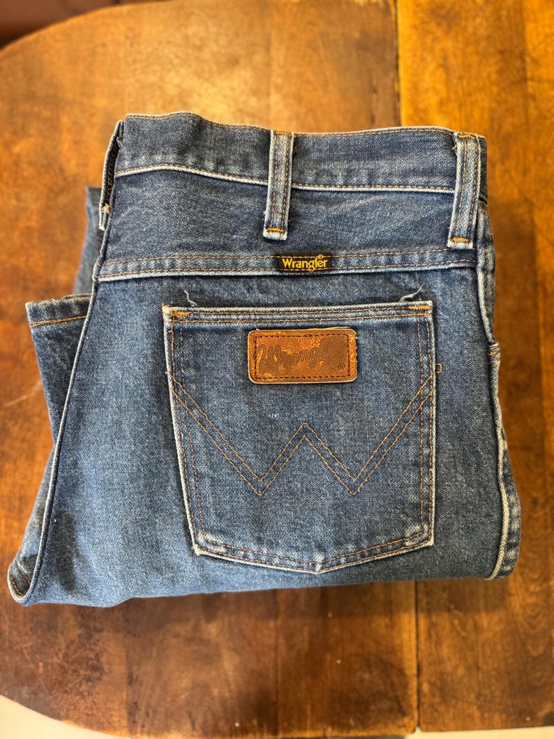 Puede incluir: Un par de pantalones vaqueros azules de mezclilla con un parche de cuero marr&oacute;n que dice "Wrangler" en el bolsillo trasero.