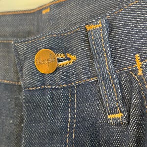 May include: Close-up of the waistband of a pair of blue denim jeans with a brown leather patch that says "Wrangler" on it.