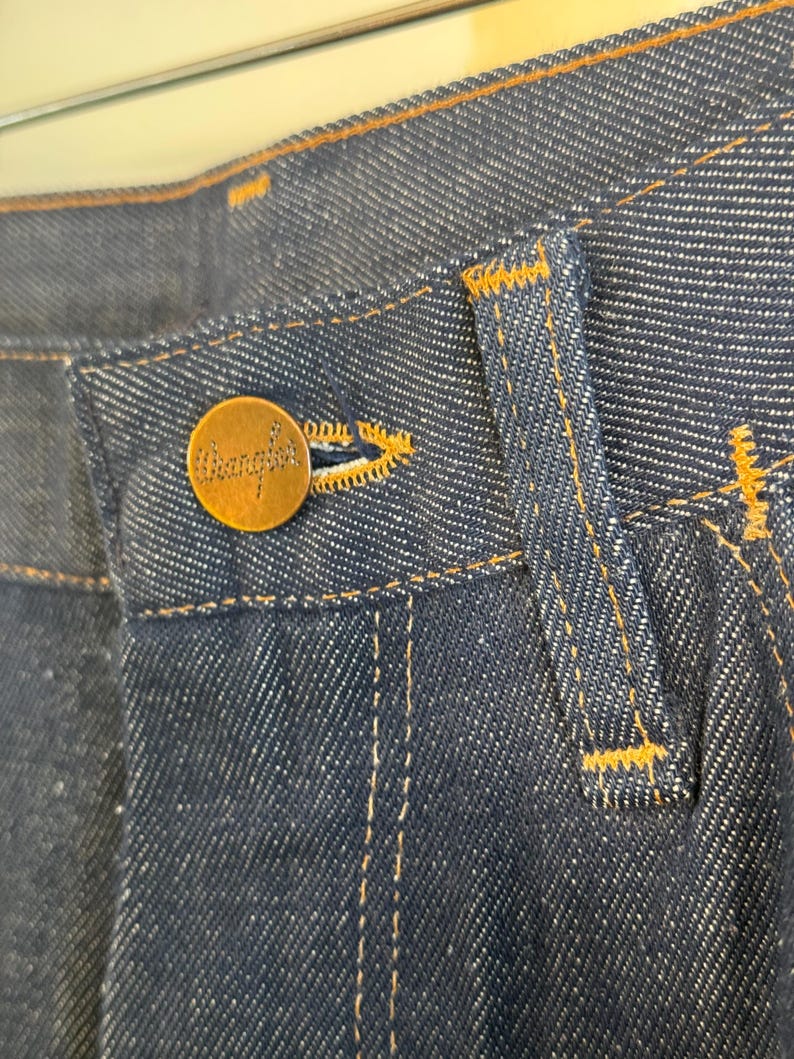 May include: Close-up of the waistband of a pair of blue denim jeans with a brown leather patch that says "Wrangler" on it.