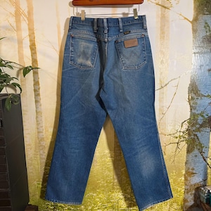 Pu&ograve; includere: Un paio di jeans blu in denim con una toppa in pelle marrone sulla tasca posteriore. I jeans sono appesi a un appendiabiti in legno.