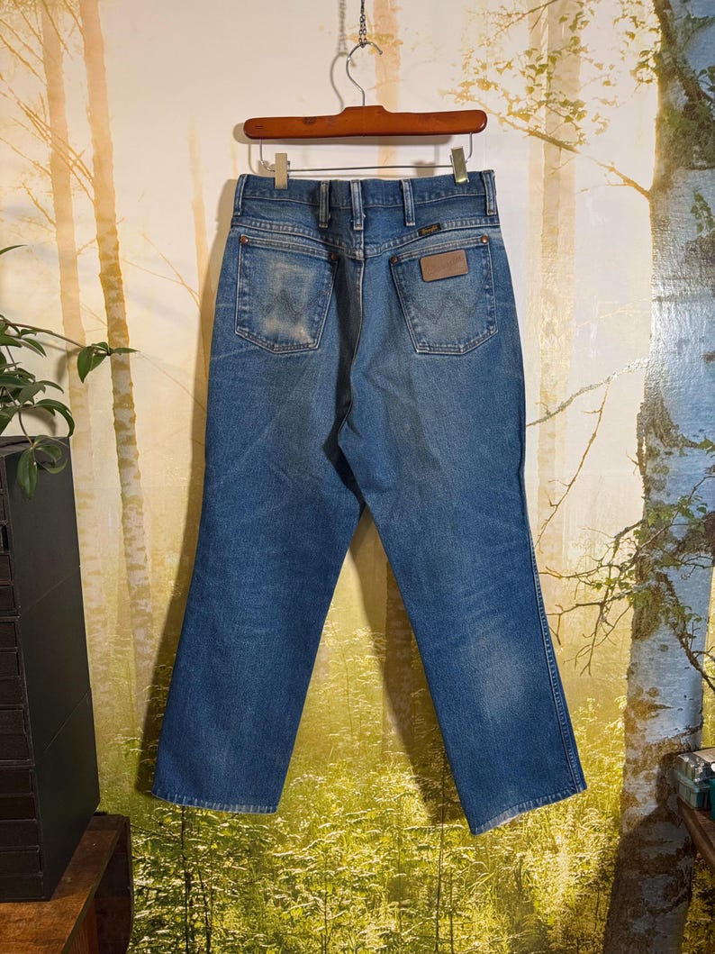 Pu&ograve; includere: Un paio di jeans blu in denim con una toppa in pelle marrone sulla tasca posteriore. I jeans sono appesi a un appendiabiti in legno.