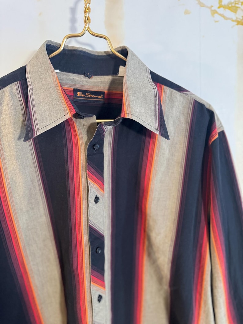 May include: A long-sleeved button-down shirt with a striped pattern in shades of black, red, orange, and gray. The shirt has a collar and a label that reads "Ben Sherman".