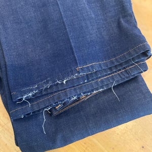 May include: A pair of dark blue denim jeans folded in half, showing the raw edge of the hem and the stitching along the seams.