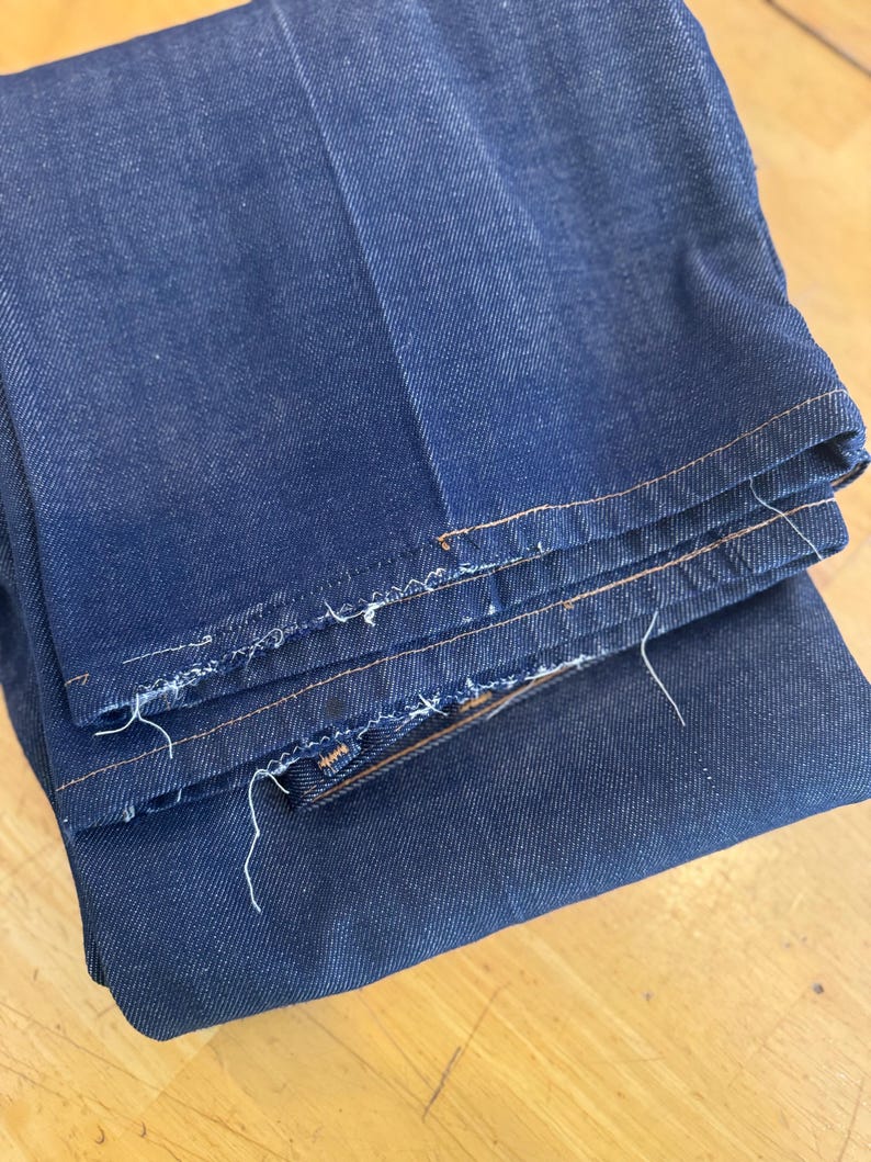May include: A pair of dark blue denim jeans folded in half, showing the raw edge of the hem and the stitching along the seams.
