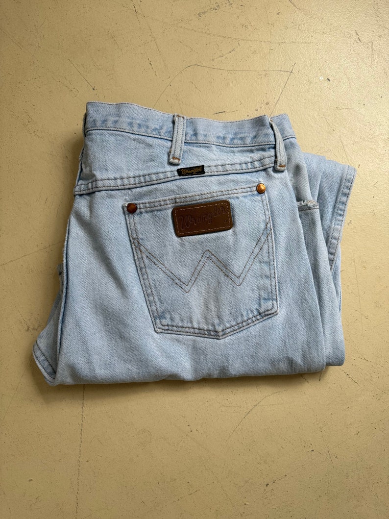 Puede incluir: Un par de pantalones vaqueros de mezclilla azul claro con un parche de cuero marr&oacute;n Wrangler en el bolsillo trasero.