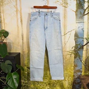May include: Light blue denim jeans hanging on a wooden hanger against a blurred background of trees. The jeans have a high waist and a straight leg.