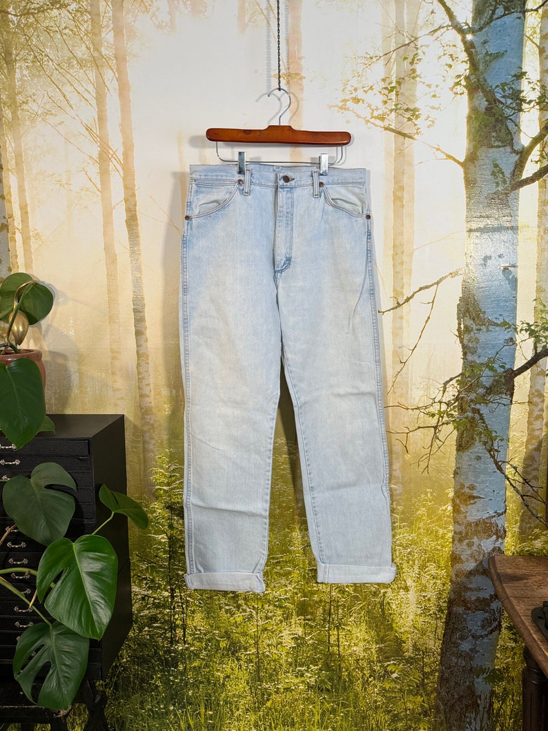 May include: Light blue denim jeans hanging on a wooden hanger against a blurred background of trees. The jeans have a high waist and a straight leg.