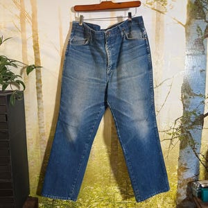 Pu&ograve; includere: Un paio di jeans blu denim con chiusura a bottone e cinque tasche. I jeans sono appesi a un appendiabiti di legno su uno sfondo di una scena di bosco.