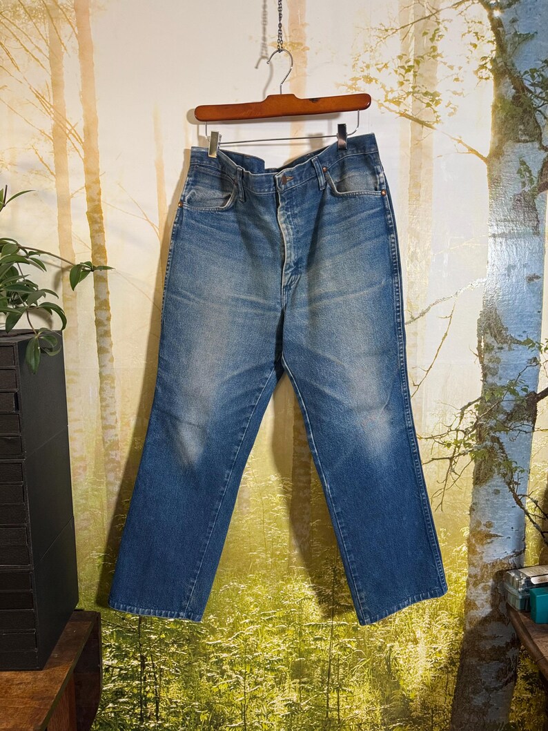 Pu&ograve; includere: Un paio di jeans blu denim con chiusura a bottone e cinque tasche. I jeans sono appesi a un appendiabiti di legno su uno sfondo di una scena di bosco.