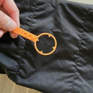 May include: Close-up of a black jacket with a small hole in the fabric. A yellow plastic tool with the word "LOOK" printed on it is used to highlight the hole.