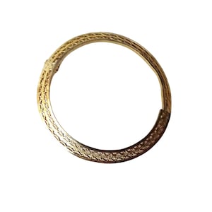 May include: A circular gold-toned metal bangle bracelet featuring a continuous engraved pattern along its entire surface. The design consists of repeating small, textured geometric marks that create a subtle, rhythmic visual detail around the band.