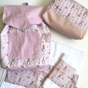 Può includere: Uno zaino rosa e bianco a quadri e con stampa floreale, una trousse da trucco rosa e bianco con stampa floreale e due asciugamani per le mani rosa e bianco con stampa floreale con il nome "Mia" ricamato.