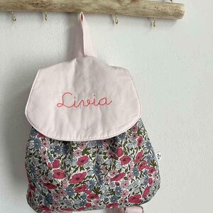 Può includere: Un piccolo zaino rosa con un motivo floreale e il nome "Livia" ricamato in rosso. Lo zaino ha spallacci regolabili e una chiusura a patta. È appeso a un attaccapanni di legno.