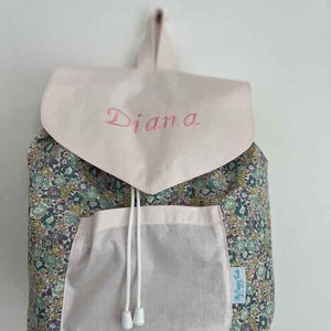 Può includere: Uno zaino rosa floreale con chiusura a coulisse bianca e una grande tasca frontale. Lo zaino ha una patta rosa con il nome "Diana" ricamato in rosa sul davanti.