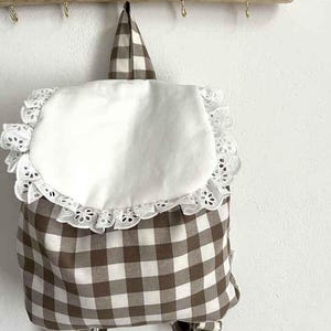 Può includere: Uno zaino a quadri marrone e bianco con una patta bianca bordata di pizzo smerlato. Lo zaino ha spallacci regolabili e una maniglia superiore. La borsa è appesa a un'asta di legno con piccoli ganci.