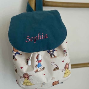 Può includere: Uno zaino in tela blu turchese e bianco con chiusura a coulisse. Lo zaino ha un nome personalizzato, "Sophia", ricamato sulla patta anteriore. Il tessuto principale presenta un motivo ripetuto di Belle e la Bestia de La Bella e la Bestia.
