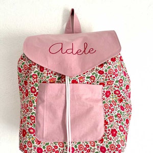 Può includere: Uno zaino rosa e floreale con il nome "Adele" ricamato sulla patta. Lo zaino presenta una tasca frontale e una chiusura con coulisse. Il motivo floreale comprende fiori rossi e foglie verdi.