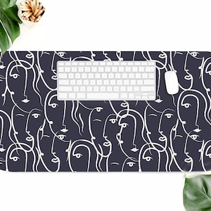 May include: A black desk mat with a white abstract pattern of faces. A white keyboard and a white computer mouse are on top of the mat.