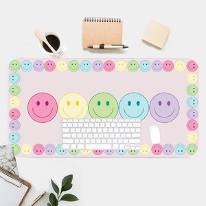 May include: A pink and white desk mat with a border of colorful smiley faces. The mat features a row of five larger smiley faces in different colors, a keyboard, and a white mouse.
