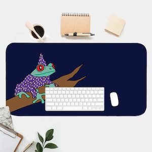 May include: A blue and purple frog wearing a wizard hat sits on a brown branch. The frog has red eyes and is sitting on a dark blue background. A white keyboard and a white mouse are on the desk.