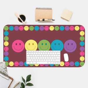 May include: A maroon desk mat with a border of colorful smiley faces. The mat features five larger smiley faces in different colors, including pink, yellow, green, blue, and purple. A white keyboard and a white mouse are on the mat.