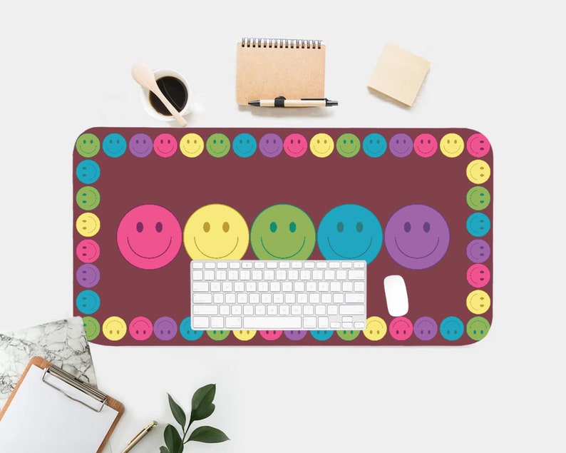 May include: A maroon desk mat with a border of colorful smiley faces. The mat features five larger smiley faces in different colors, including pink, yellow, green, blue, and purple. A white keyboard and a white mouse are on the mat.