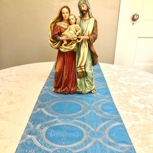 May include: A blue table runner with a silver pattern of circles and crosses. A figurine of the Holy Family is placed on top of the runner.