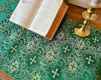 May include: A green altar cloth with a gold and silver pattern of crosses and floral designs. A small open book and a gold candlestick are on the cloth.