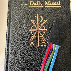 K&ouml;nnte beinhalten: Ein schwarzes, ledergebundenes Buch mit goldenen Lettern, auf dem "Saint Joseph Daily Missal" steht. Das Buch hat ein goldenes Symbol auf dem vorderen Umschlag und ein schwarzes Lederlesezeichen mit bunten B&auml;ndern.