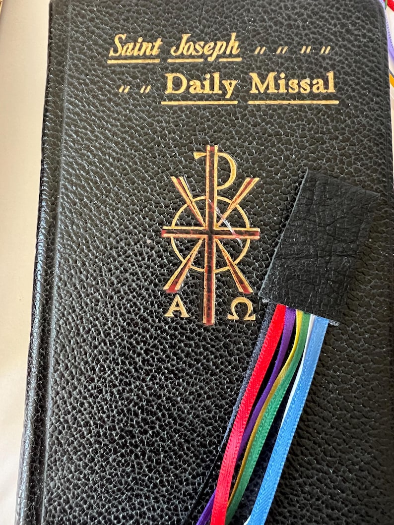 K&ouml;nnte beinhalten: Ein schwarzes, ledergebundenes Buch mit goldenen Lettern, auf dem "Saint Joseph Daily Missal" steht. Das Buch hat ein goldenes Symbol auf dem vorderen Umschlag und ein schwarzes Lederlesezeichen mit bunten B&auml;ndern.