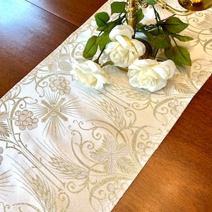May include: A white table runner with a gold embroidered design of wheat, grapes, and a cross. The runner is decorated with white roses and a gold candelabra.